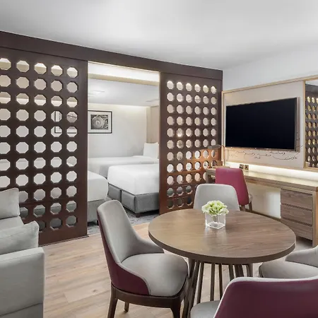 Makarem Ajyad Makkah Hotel Mecca Exterior photo The photo shows a modern hotel room or suite. It features a living area with a comfortable gray sofa and a round wooden dining table surrounded by upholstered chairs. A large flat-screen TV is mounted on the wall opposite the seating area. There's a 