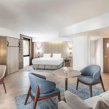 Makarem Ajyad Makkah Hotel Mecca Exterior photo The photo shows a modern hotel room with a spacious layout. It features a large bed with a simple headboard against a light-colored wall. There are two soft chairs in light blue and beige positioned around a small round table. The flooring is wooden,